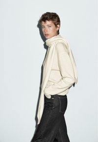 Donna con capelli corti indossa un maglione crema e una sciarpa, jeans neri, in piedi di lato con le mani nelle tasche su uno sfondo chiaro uniforme.