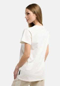 Cremefarbendes T-Shirt mit kurzen Ärmeln aus weicher Baumwolle. Verfügt über einen runden Ausschnitt und Seitenschlitze mit einem schwarzen Akzent-Label am Saum.