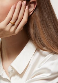 Gold ring with a wavy design, set against a light background. Hand rests near the face, showcasing natural nails and a silk blouse.