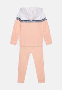 Világos rózsaszín tréningruha, fehér kapucnival, bordás mandzsettával és tengerészkék szalaggal, rajta a "LOVE GUESS" logóval. Sima tapintású, testhezálló kialakítás.