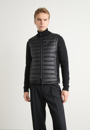 Black puffer vest featuring a zip front, high collar, and quilted design. Worn over a black sweater, paired with black trousers.