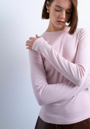 Woman with short brown hair wears light pink long-sleeve shirt and gold earrings, looking down with one arm crossed and hand touching opposite shoulder.