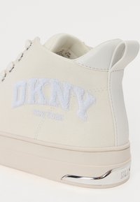 Balti kanvoso sportbačiai su iškiliu "DKNY" logotipu, odiniais akcentais ir metaliniais detalėmis ant kulno. Turi apvalias raišteles ir lygų paviršių.