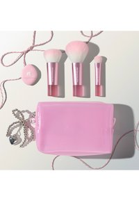 Drie roze make-upkwasten, een roze spons en een kleine roze cosmeticatasje met een zilveren bedel op een wit oppervlak.