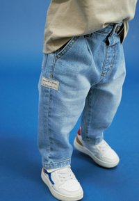 Jeans in denim azzurro con taglio relaxed fit, caratterizzati da un'etichetta con la scritta "AUTHENTIC GOODS" sulla tasca, abbinati a sneakers bianche.