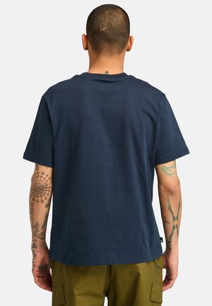 Marineblaues Baumwoll-T-Shirt mit Rundhalsausschnitt und kurzen Ärmeln, das eine schlichte Textur und eine lockere Passform aufweist. Sichtbare Tätowierungen an den Armen.