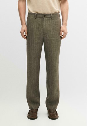 Mand iført olivengrønne bukser med nålestriber og brune loafers, stående mod en ensartet lys baggrund.