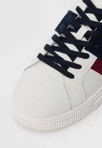 Sneaker featuring a white textured leather upper, navy suede accents, round navy laces, and a white rubber sole with ribbed detailing.