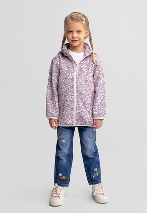 Jauna meitene ar pītu matu sakārtojumu, vilkusi ziedu raksta rāvējslēdzēja jaku, izšūtas zilās džinsu bikses un baltas sporta kurpes, stāv pret vienkāršu fonu.