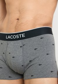 Gray cotton boxer shorts with small black Lacoste logos, featuring a black waistband with white "LACOSTE" lettering. Smooth texture.