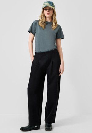 Frau trägt ein ausgebleichtes grünes T-Shirt, schwarze weite Hosen, schwarze Stiefel und eine blau-grüne Batik-Basecap und steht vor einem schlichten Hintergrund.
