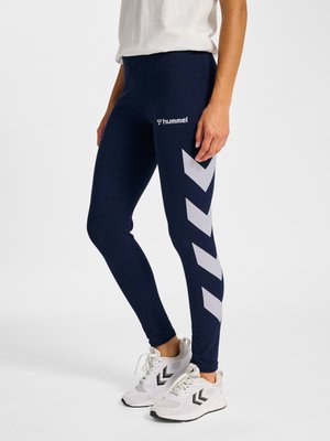 Persona que lleva leggings azul marino con franjas en zigzag blancas y el logo "hummel", combinados con zapatillas blancas con zigzags negros.