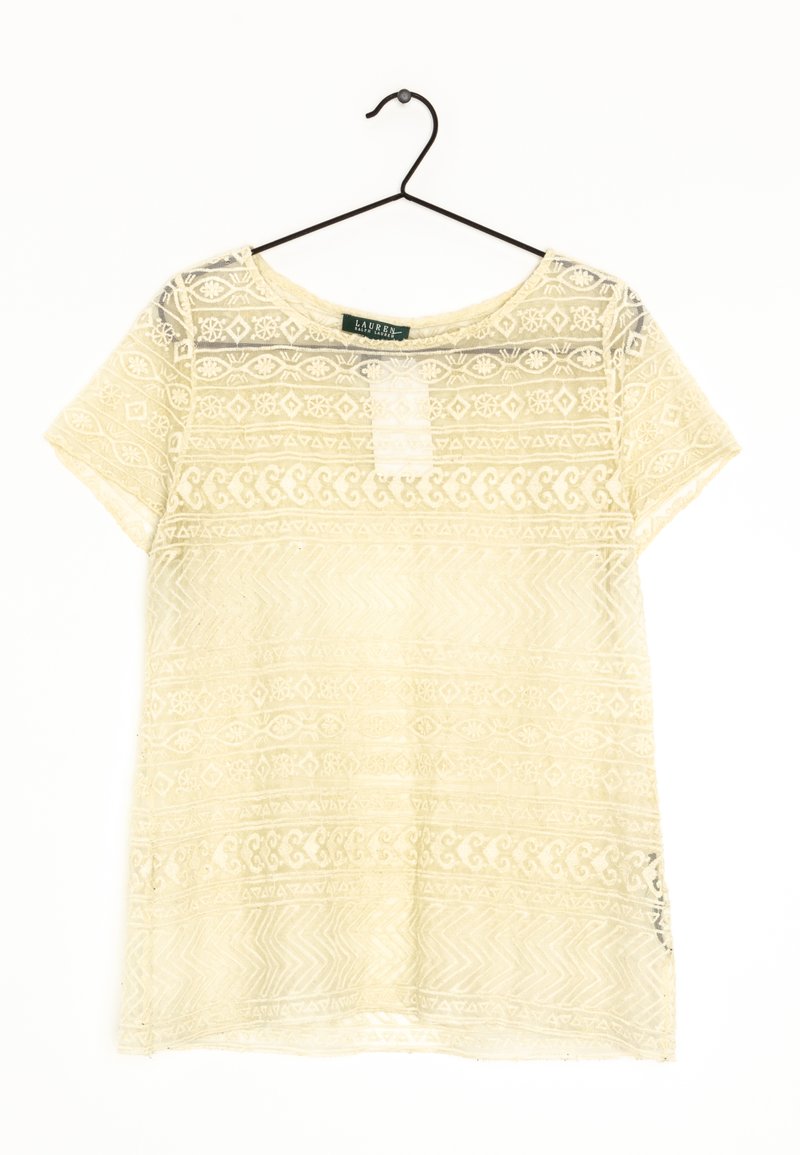 Kortärmad crèmelace-topp med blommönster och geometriska mönster, hängande på en svart trådhängare mot en vit bakgrund.