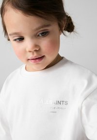 Balta medvilnės džemperis su metaliniu "ALLSAINTS" logotipu ir tekstu įvairiomis kalbomis. Dizainas turi apvalų iškirpimą ir trumpas rankoves.