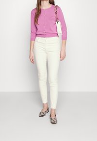 Cárdigan rosa claro con mangas largas y botones, combinado con jeans ajustados blancos y zapatos planos de estampado de serpiente blanco y negro.