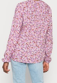 Blouse florale violette à manches longues, avec un détail froncé à l'arrière. Tissu léger et doux avec un mélange de fleurs blanches, jaunes et rouges.