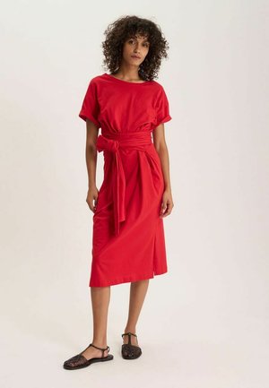 Femme aux cheveux bouclés portant une robe midi rouge avec un lien à la taille et des sandales plates noires tressées, debout devant un fond uni.