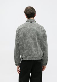 Chaqueta de denim gris con un patrón abstracto, puños abotonados y un cuello. La parte trasera presenta un diseño texturizado sutil. Lleva puesta con pantalones negros.