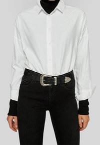 White button-up shirt with long sleeves, paired with a black belt featuring a silver buckle, worn over black jeans.