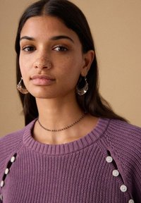 Pull tricoté violet avec col côtelé et détails de boutons sur le côté. Le modèle porte des boucles d'oreilles anneaux argentées et un collier de perles. Fond neutre.