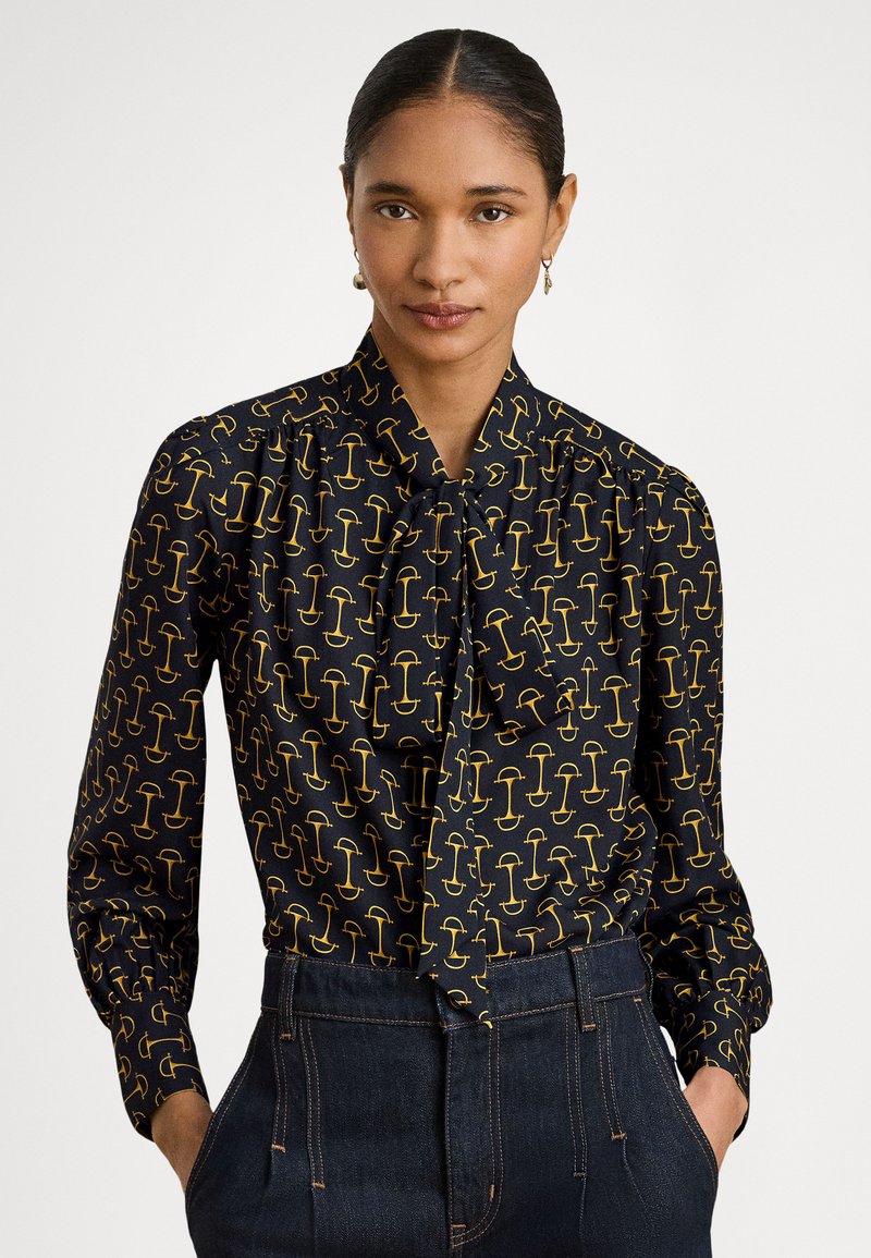 Crna bluza sa žutim apstraktnim uzorkom, dugim rukavima, ovratnikom sa mašnom i teksturiranim tkanjem. Kombinira se s tamnim trapericama visokog struka.