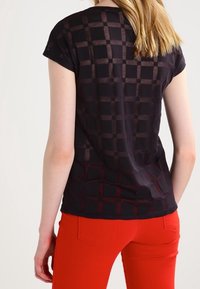 Černý top s kostkovaným vzorem, krátkými rukávy a měkkou, průsvitnou texturou. V kombinaci s jasně červenými kalhotami s zadními kapsami.