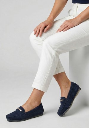 Kvinde iført hvide bukser og mørkeblå ruskindslædermokkasiner, siddende med hænder hvilende på sit knæ mod en ensartet baggrund.