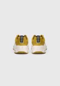 Tênis atléticos amarelos e brancos com sola texturizada, colarinho acolchoado e uma alça preta com detalhes brancos.