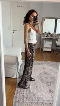 Mulher com cabelo encaracolado a usar uma t-shirt branca e calças de padrão de leopardo tira uma selfie ao espelho numa sala com chão de madeira e um tapete estampado.