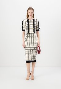 Rochie bej până la genunchi, cu un model de grilă neagră, mâneci puff texturate, detalii cu butoane aurii și un tiv negru, asortată cu o geantă roșie mică.