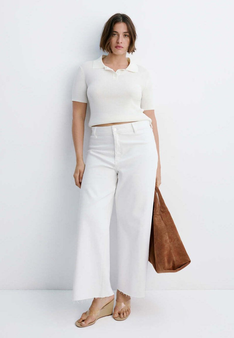 Femme aux cheveux courts et bruns portant un haut blanc à manches courtes et un pantalon blanc à jambes larges, tenant un grand sac marron, debout contre un mur blanc.