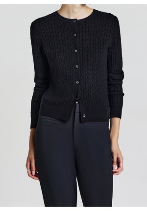 Femme portant un cardigan noir cintré en maille torsadée boutonné, associé à un pantalon noir taille haute ajusté, sur un fond uni.