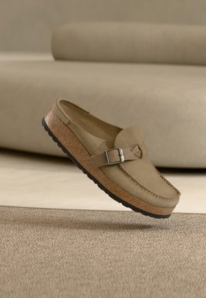 Beige suede loafer with adjustable buckle strap, cork sole, and black tread, positioned above a matching textured carpet.