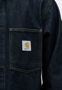 Σκούρο τζιν μπουφάν με εμφανές μπροστινό τσεπάκι που διαθέτει ραφές σε απόχρωση καμηλοπάρδαλης και λευκή ραφή λογότυπου Carhartt.