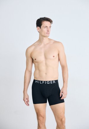 Shirtloze man met kort bruin haar, gekleed in zwarte Hilfiger boxershorts, staand tegen een effen lichtgrijze achtergrond, kijkend naar rechts.