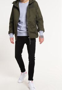 Olivengrøn hættejakke med lynlås, grå sweatshirt under, sorte bukser og hvide sneakers med røde accenter. Har sidelomme.