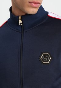 Chaqueta de punto azul marino con un cuello alto, que presenta acentos rojos en los hombros y un detalle de logo hexagonal dorado en el pecho. Textura de tejido suave.