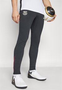Pantaloni sportivi neri con texture a coste, logo del FC Barcelona, accenti rossi sui lati, abbinati a scarpe da calcio bianche e a una palla con motivo.
