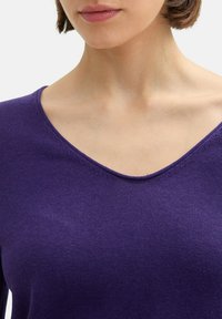V-Ausschnitt-Oberteil in tiefem Lila, aus weichem, dehnbarem Stoff. Glatte Textur mit schmalem Schnitt, der den Ausschnitt und die Schultern betont.