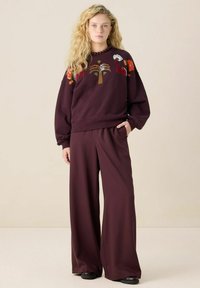 Vrouw met lang stijl blond haar die een bordeauxrode geborduurde sweater en wijde broek draagt, staand met één hand in de zak.