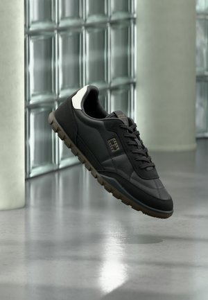 Schwarzer Sneaker mit weißer Fersenlasche und strukturierter brauner Sohle, der in Innenräumen nahe einer Glassteinwand und einem Betonboden schwebt.