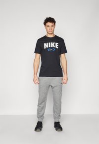 Crna pamučna majica s bijelim i plavim "NIKE" logotipom, u kombinaciji sa sivim trenerkama i crnim sportskim cipelama. Jednostavan dizajn, ležeran kroj.
