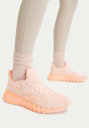 Beine, die beige Leggings, hellrosa gerippte Socken und pastellpfirsichfarbene Sportschuhe mit profilierten Sohlen auf weißem Hintergrund tragen.