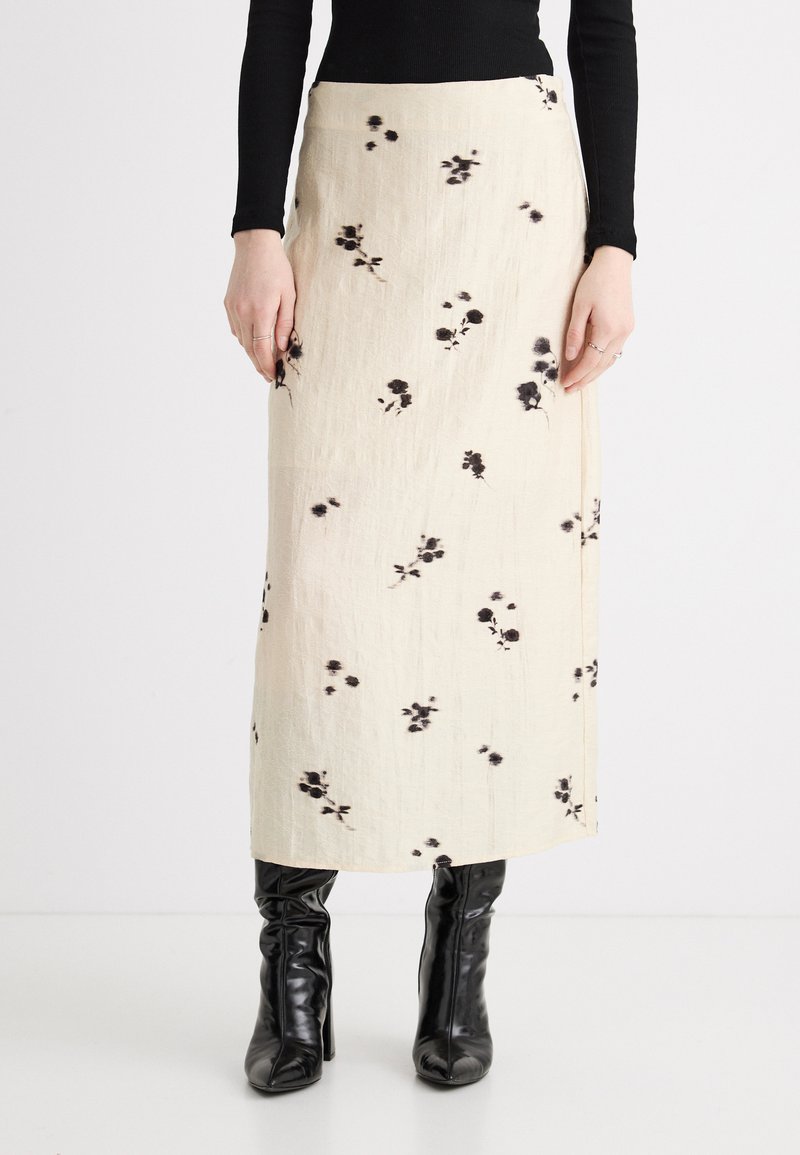 Κρέμ midi φούστα με μαύρο λουλουδάτο σχέδιο, συνδυασμένη με μαύρο μακρυμάνικο τοπ και μαύρες γυαλιστερές μπότες με τακούνι, εμφανιζόμενη από τον λαιμό μέχρι τα πόδια.