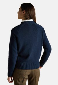 Tumes sinine kudumiga sviiter ribatud ääre ja varrukatega, millel on tõstetud krae ja õmblusteta õlavarre disain. Kantakse valge särgi kohal.