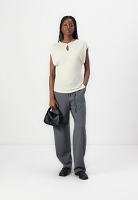 Haut beige clair à manches courtes avec un décolleté en forme de clé, associé à un pantalon gris ample. La modèle tient un petit sac à main noir.