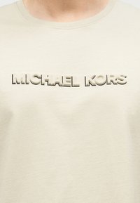 Μπεζ βαμβακερό μπλουζάκι με ανάγλυφο, τονισμένο λογότυπο "MICHAEL KORS" σε αντίθετο χρώμα στο στήθος. Κλασικός στρογγυλός λαιμός, κοντά μανίκια.