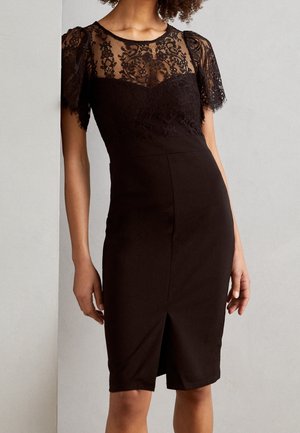 Woman wearing a black knee-length dress with sheer lace short sleeves and lace bodice, standing against a plain light wall background.