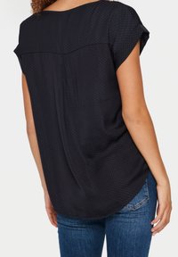 Blouse noire à manches courtes avec un motif à pois, coupe décontractée, dos plissé et ourlet arrondi, associée à un jean bleu.