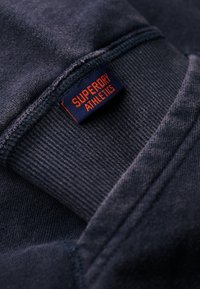 Close-up van een marineblauwe sweatshirtmanchet met geribbelde textuur en een klein marineblauw label met de oranje tekst "SUPERDRY ATHLETICS".